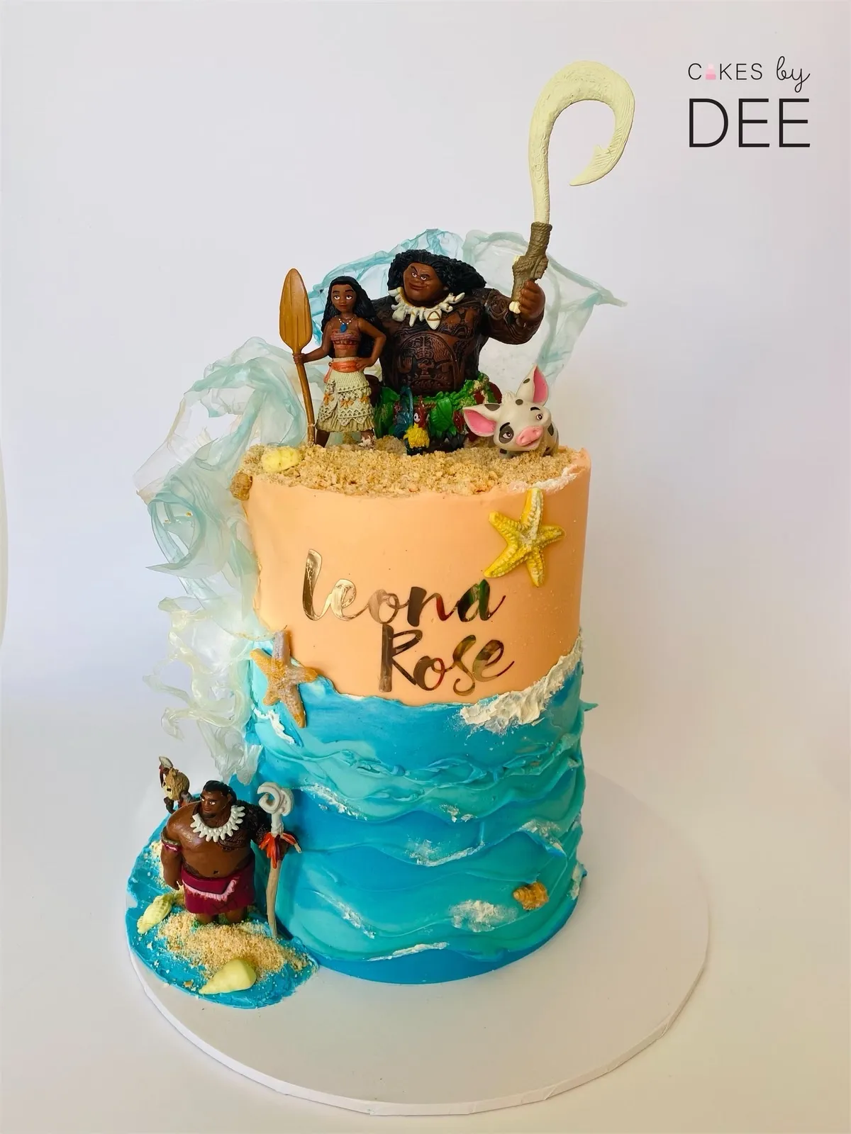 Two-tier Moana-themed birthday cake with ocean wave buttercream, character figurines and Leona Rose gold lettering