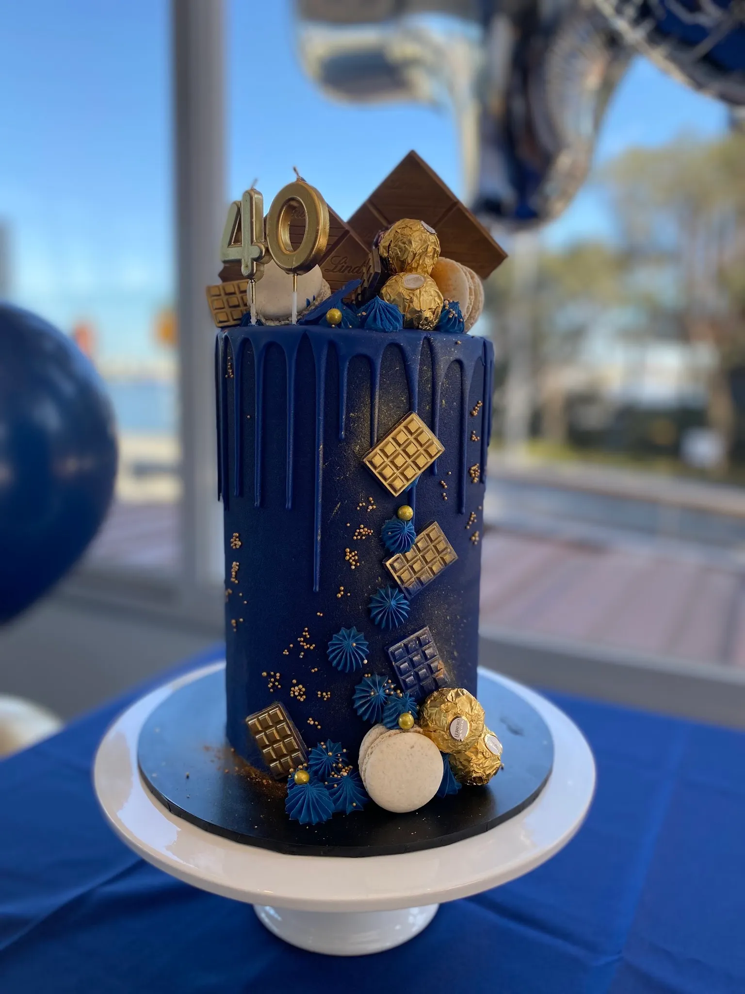 Tall navy blue drip cake with gold chocolates, macarons and Ferrero Rocher on a white stand with waterfront window behind