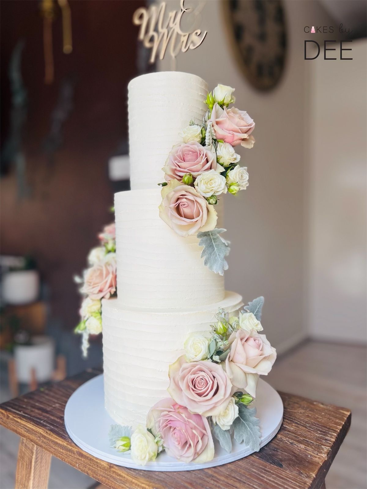 Four-tier white ribbed wedding cake with cascading fresh pink roses and Mr and Mrs gold topper on a wooden stand