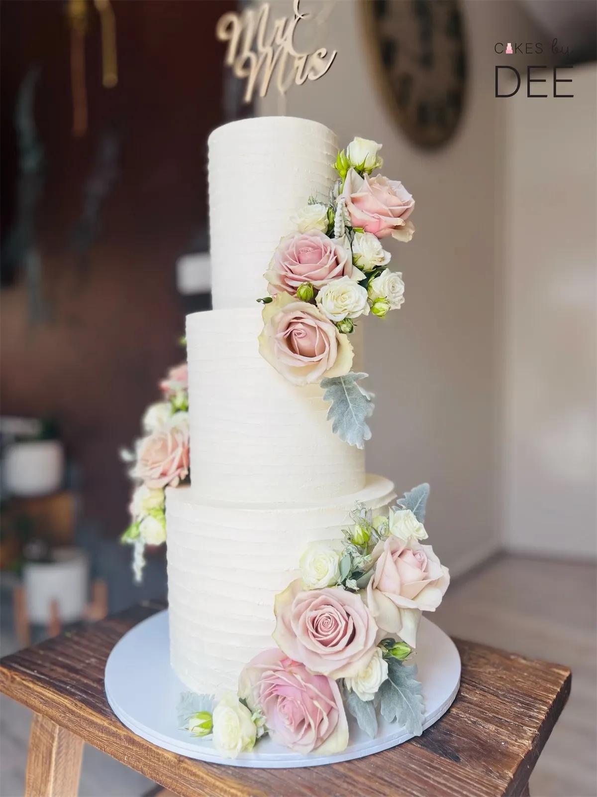 Four-tier white ribbed wedding cake with cascading fresh pink roses and Mr and Mrs gold topper on a wooden stand