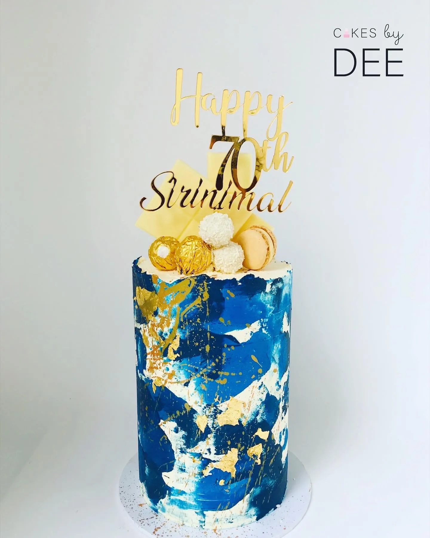 Cylindrical abstract-painted blue and gold birthday cake with macarons and Happy 70th Sirinimal gold topper