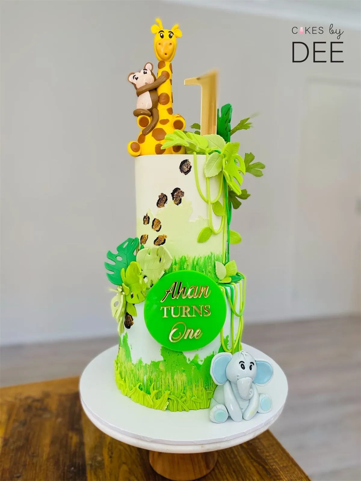 Two-tier jungle safari 1st birthday cake with giraffe, monkey and elephant figurines and tropical leaf decorations