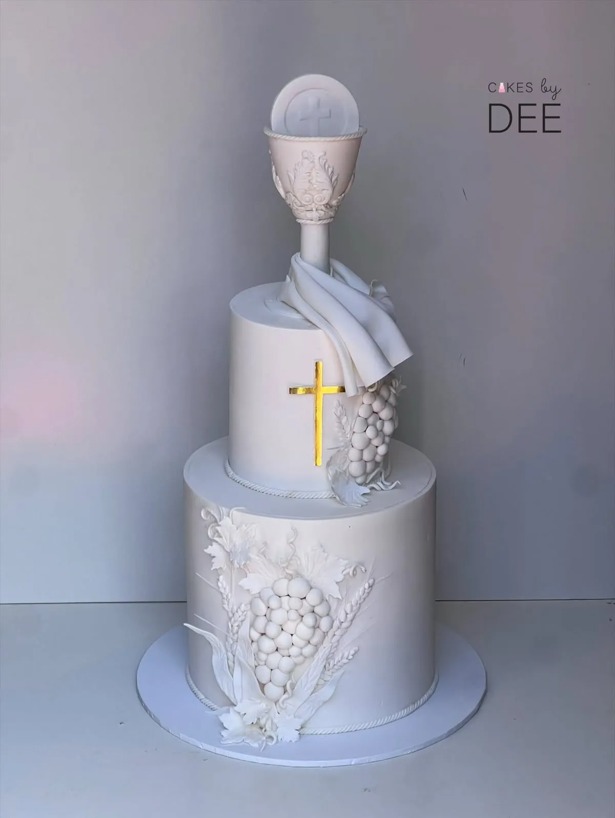 Full view of two-tier white fondant First Communion cake with sculpted grapes, gold cross and ornate chalice topper